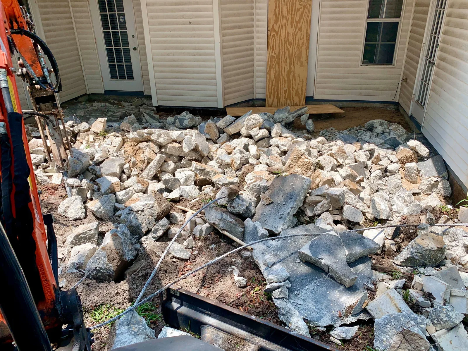 Concrete patio broken into slab pieces behind a house, with an excavator boom at the edge of the frame ready to load the rubble