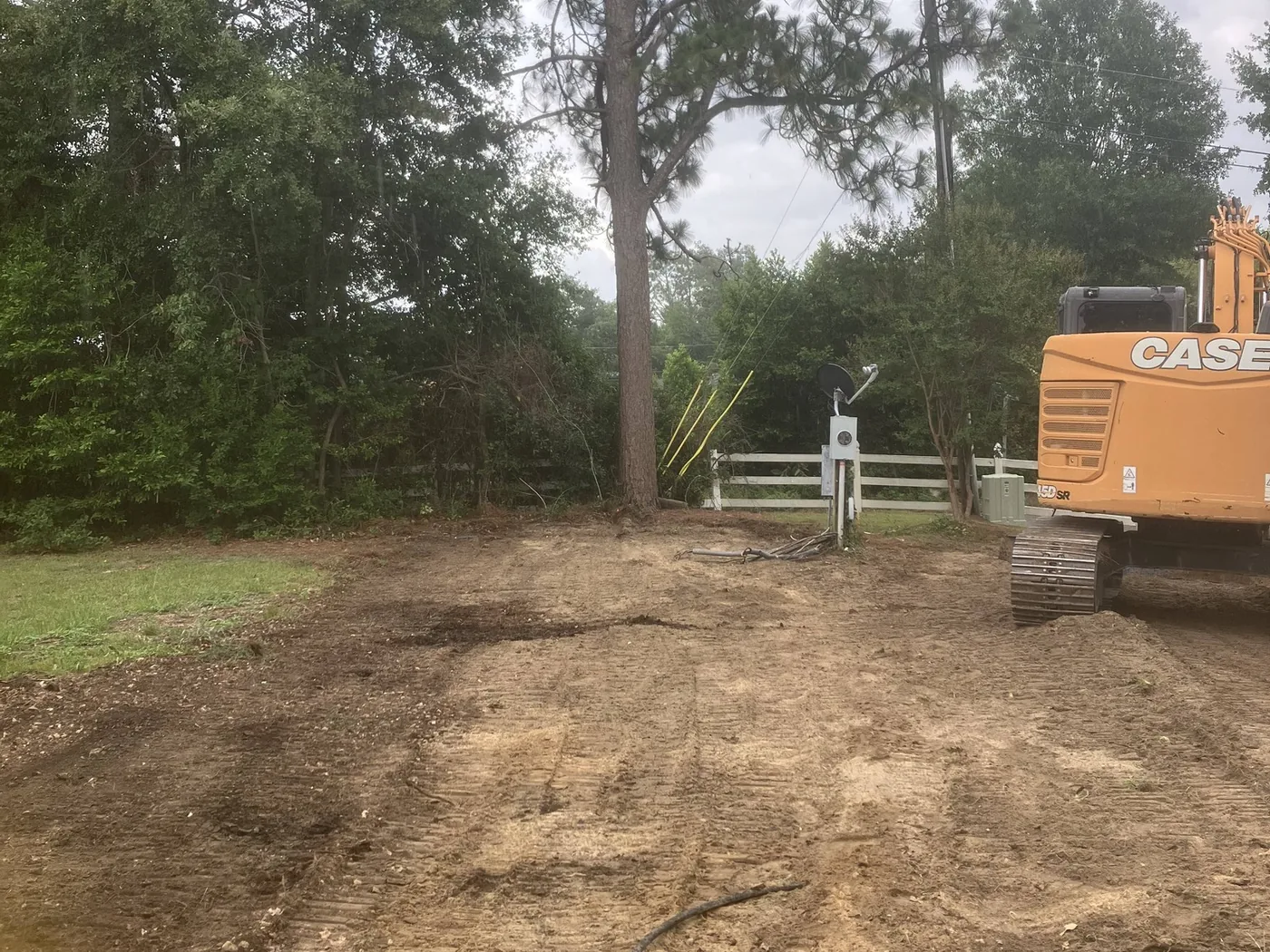 Freshly graded lot with a pipe stub and marker flag in the foreground and an excavator at the tree line