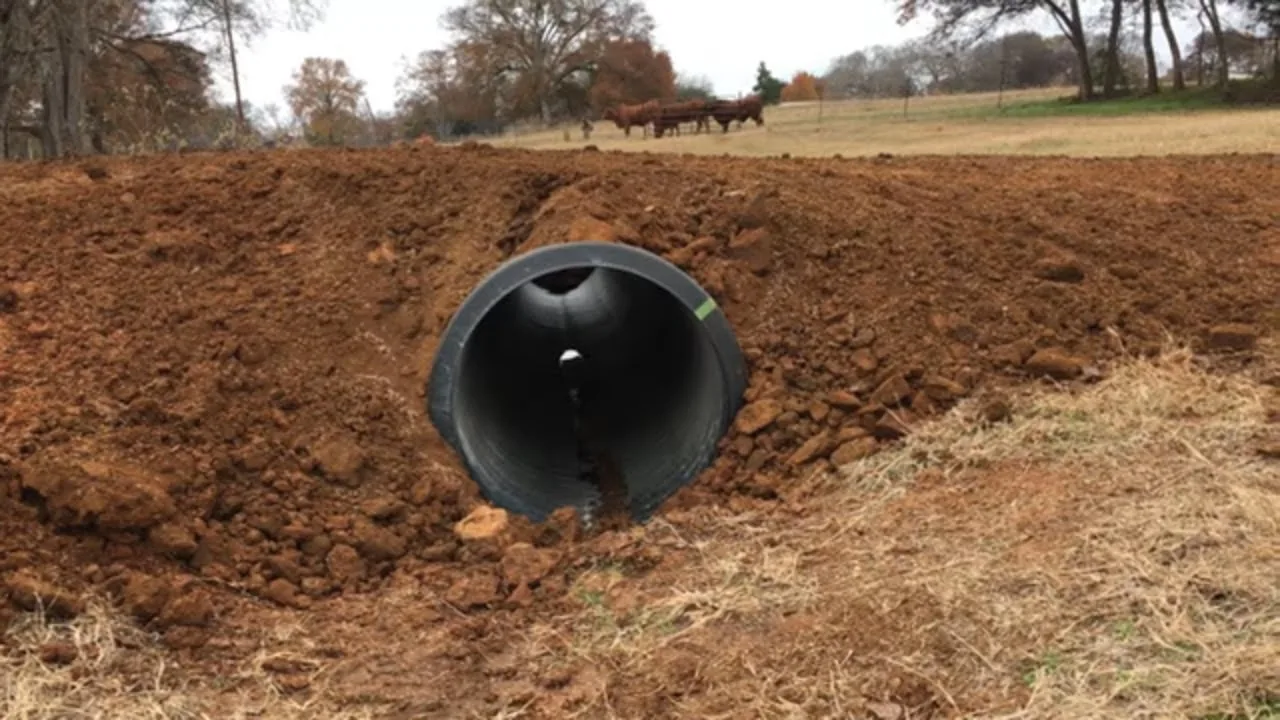 New black plastic culvert pipe set under a freshly packed red clay crossing at the edge of a pasture