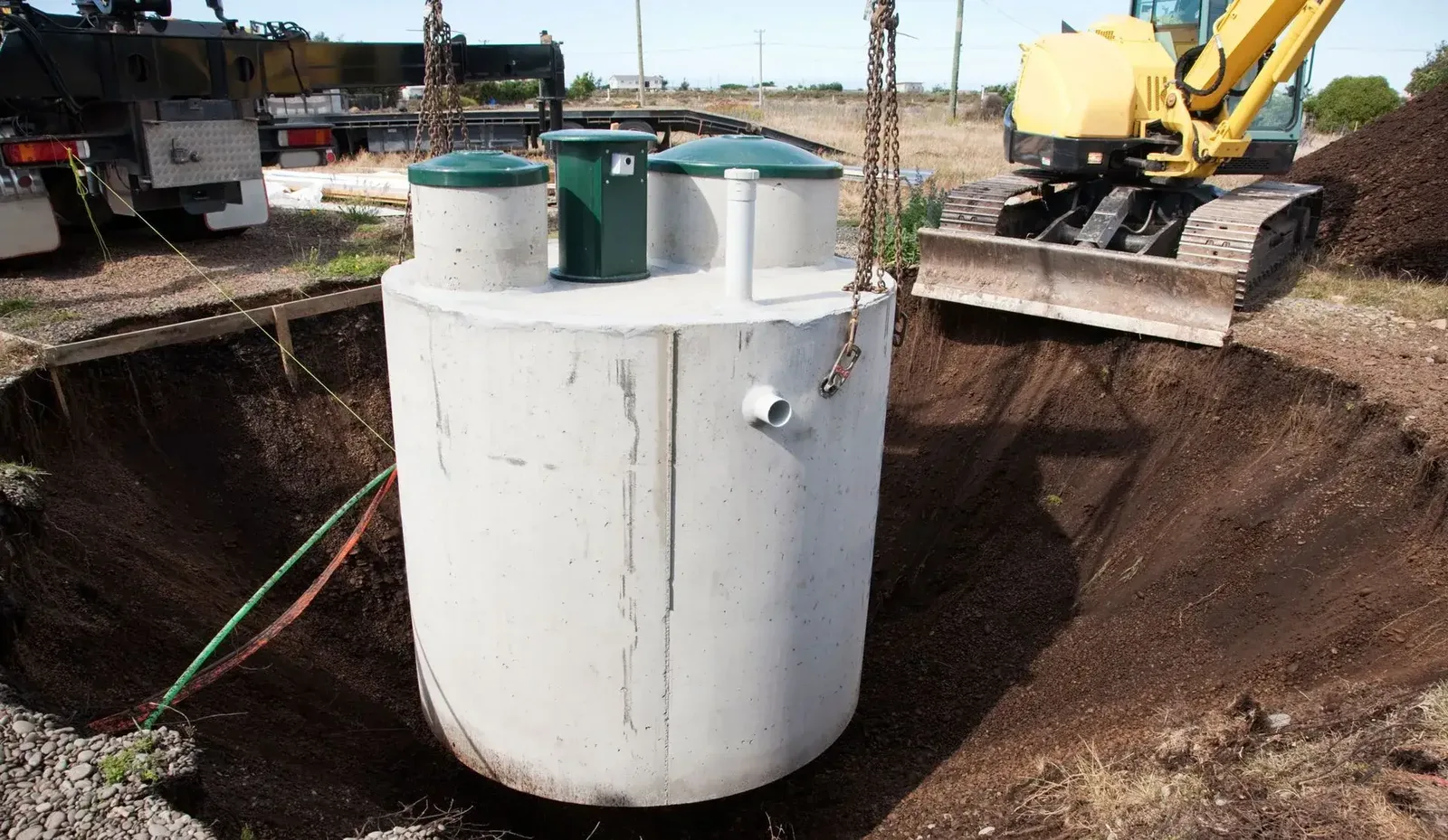 Concrete septic tank with green riser lids lowered on chains into an open excavation, with an excavator blade parked at the edge of the hole